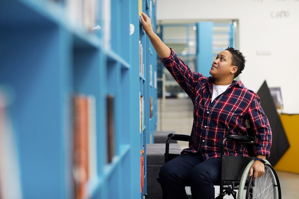 Portrait of disabled student in wheelchair choosing books while studying in college library, copy space