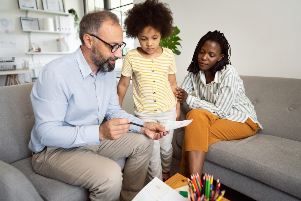 Mature adult male psychologist talking with little girl in the office about her drawing.