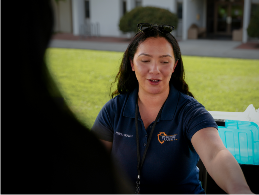 A Health Ambassador reach across a table to hand a resident a pamphlet with resources and information related to health.