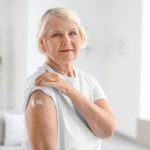 Older woman in a white shirt shows a small bandage on her upper arm after receiving a vaccination.