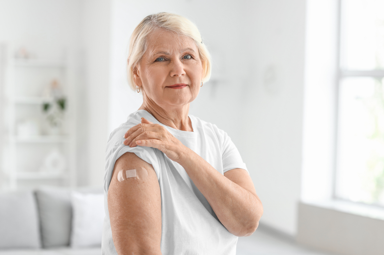 Older woman in a white shirt shows a small bandage on her upper arm after receiving a vaccination.
