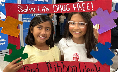 Two girls take a photo with a frame that says, "solve life drug free!" red ribbon week.