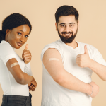 Two people smiling and giving thumbs up while showing bandages on their upper arms after getting vaccinated.