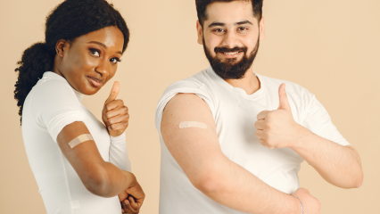 Two people smiling and giving thumbs up while showing bandages on their upper arms after getting vaccinated.
